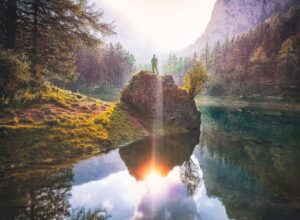 A person standing on a sunlit rock surrounded by forest and water, reflecting solitude and awareness.